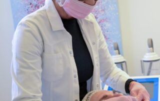 Heilpraktikerin in weißem Kittel, rosafarbenem Mundschutz und Handschuhen am Kopfende einer Patientin stehend, welche auf einer Behandlungsliege liegt