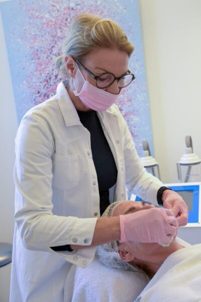 Heilpraktikerin in weißem Kittel, rosafarbenem Mundschutz und Handschuhen am Kopfende einer Patientin stehend, welche auf einer Behandlungsliege liegt